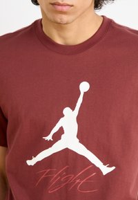 Mørkebrun bomulds t-shirt med en stor hvid silhuet af en springende figur og ordet "Flight" i rød kursiv nedenfor designet.