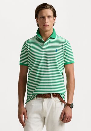 Cămașă polo cu dungi verzi și albe, având un guler verde contrastant. Prezintă un mic logo albastru pe piept. Este purtată cu pantaloni de culoare deschisă.
