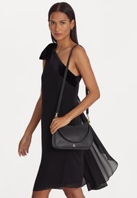 Mujer viste un vestido negro sin mangas con un detalle de terciopelo en el hombro, sosteniendo un bolso de cuero negro cruzado con herrajes dorados.