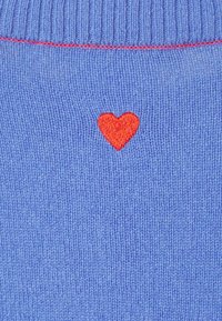 Blaue gestrickte Stoff mit einem kleinen roten gestickten Herzen nahe dem unteren Ende. Der gerippte Halsausschnitt verleiht dem Gesamtdesign Textur.