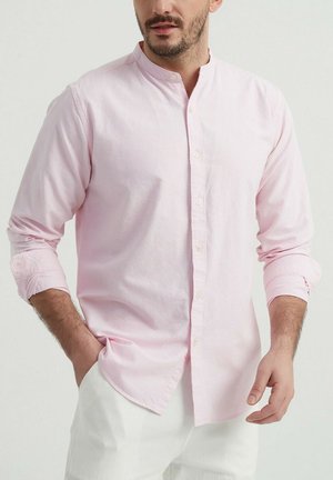 Chemise longue à manches longues de couleur rose clair avec un col corseté et une fermeture à boutons sur le devant. Présente des manches retroussées et un tissu doux et texturé.