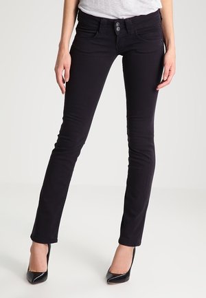 Černé úzké džíny s dvojitým zapínáním nošené ženou, kombinované s černými špičatými lodičkami a světle šedým topem.