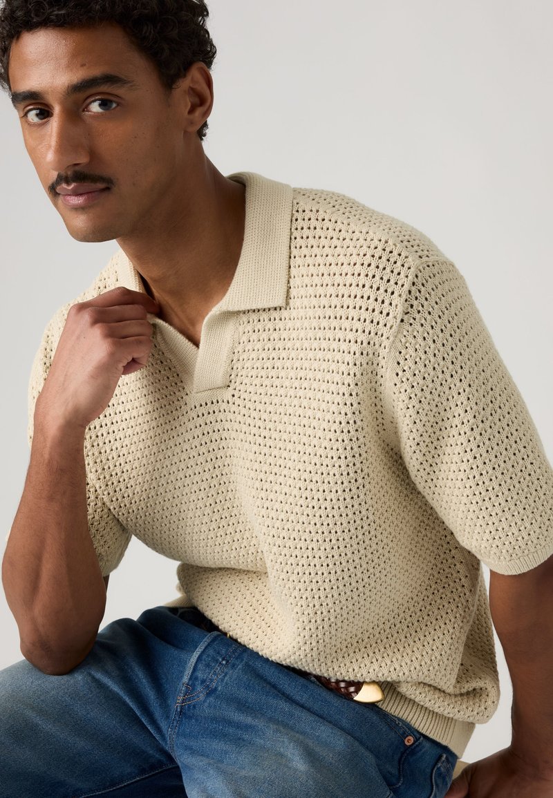 Hombre con cabello corto y rizado y bigote, lleva un polo de punto beige de manga corta y jeans azules, sentado con la mano cerca del cuello sobre un fondo liso.