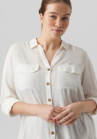 Witte, lichte blouse met een knoopsluiting aan de voorkant, twee borstzakken en oprolbare mouwen. Bevat een subtiele glans en schelpachtige knopen.