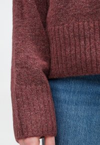 Κοντινό πλάνο ενός ατόμου που φοράει μωβ ριγωτό πλεκτό μανίκι και μπλέ τζιν, δείχνοντας λεπτομέρειες υφάσματος.