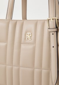 Sac à main en cuir beige avec un design matelassé horizontal, arborant un logo doré et une bandoulière robuste avec un détail de boucle.