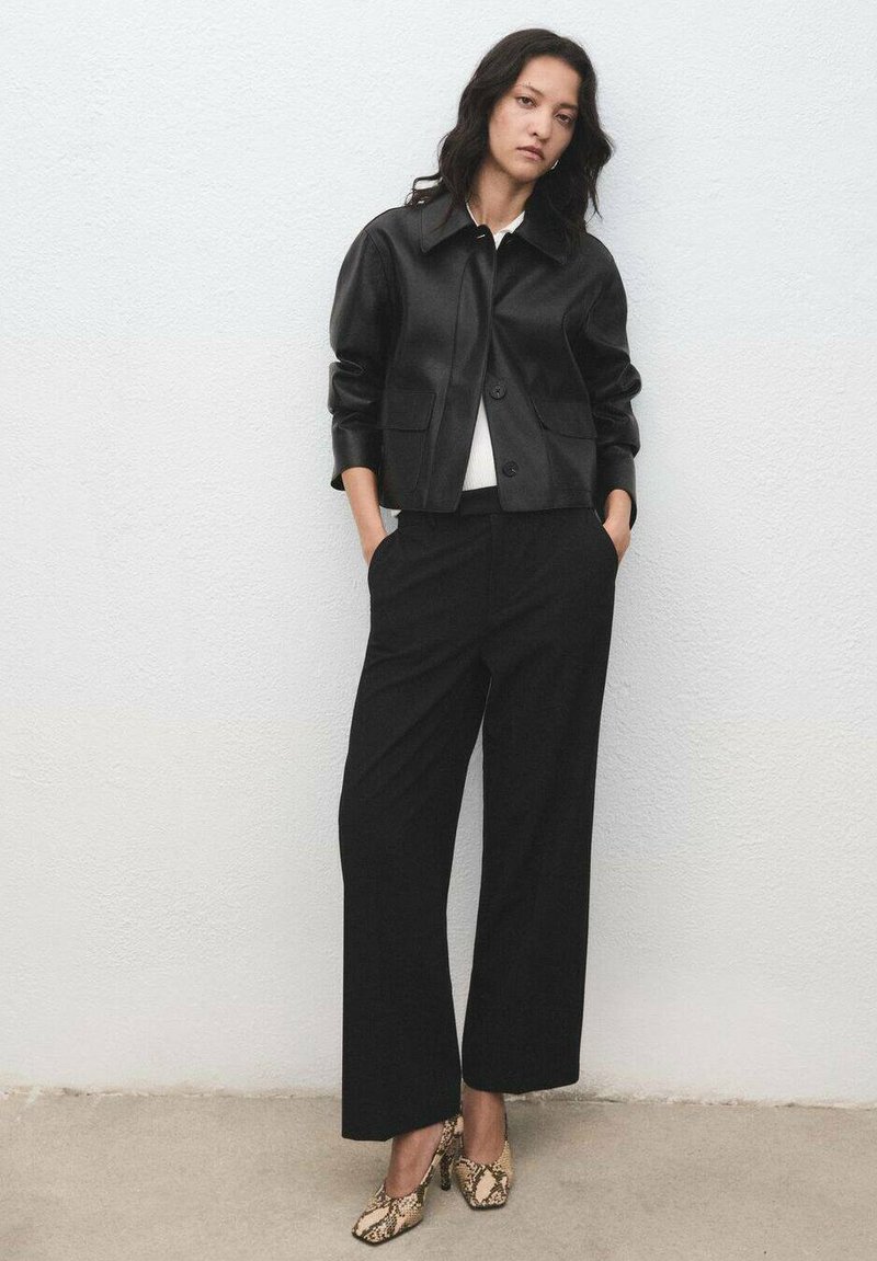 Donna che indossa una giacca di pelle nera, pantaloni larghi neri, una maglietta bianca e tacchi alti con motivo serpentino, in piedi contro un muro bianco.
