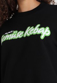 Black sweatshirt featuring bold, green letters that read "Gemüse Kebap." Fabric appears soft with a relaxed fit. Ribbed neckline visible.