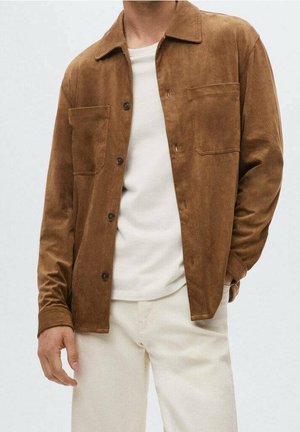 Mężczyzna w brązowej zamszowej kurtce na guziki, białym t-shircie i kremowych spodniach, stojący z jedną ręką w kieszeni.