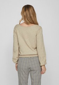 Beige Strickpullover mit weitem Ausschnitt und langen, leicht puffigen Ärmeln. Kombiniert mit karrierten Hosen, die ein kleines Rautenmuster aufweisen.