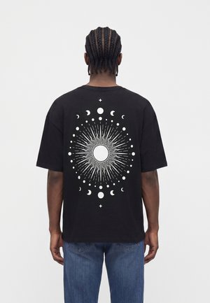 Homme aux cheveux tressés portant un t-shirt noir avec un motif circulaire blanc des phases du soleil et de la lune dans le dos, associé à un jean bleu.