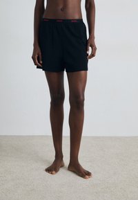 Sorte bomullsshorts med elastisk midjebånd som har rødt "HUGO" merke. Rett snitt, midt på låret lengde, og glatt tekstur.