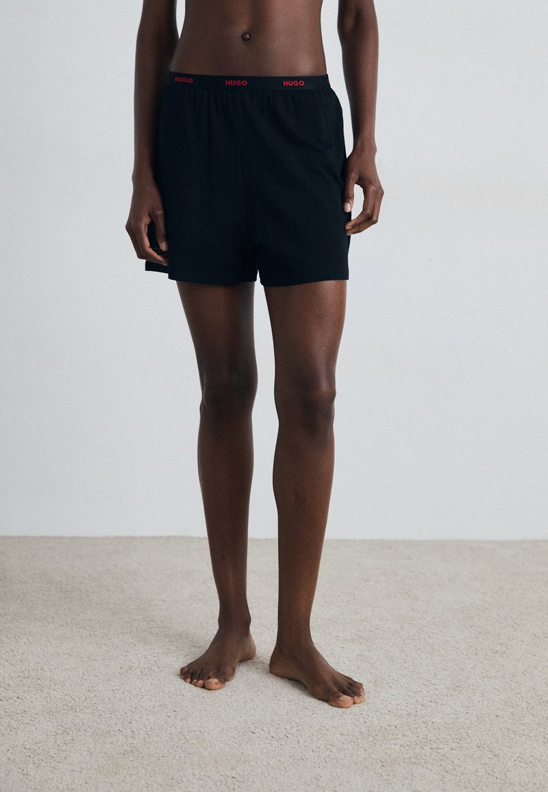 Sorte bomullsshorts med elastisk midjebånd som har rødt "HUGO" merke. Rett snitt, midt på låret lengde, og glatt tekstur.