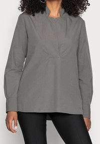 Blusa grigia a maniche lunghe con colletto alto, caratterizzata da una piega frontale e un taglio dritto. Realizzata in un tessuto liscio con una leggera lucentezza.