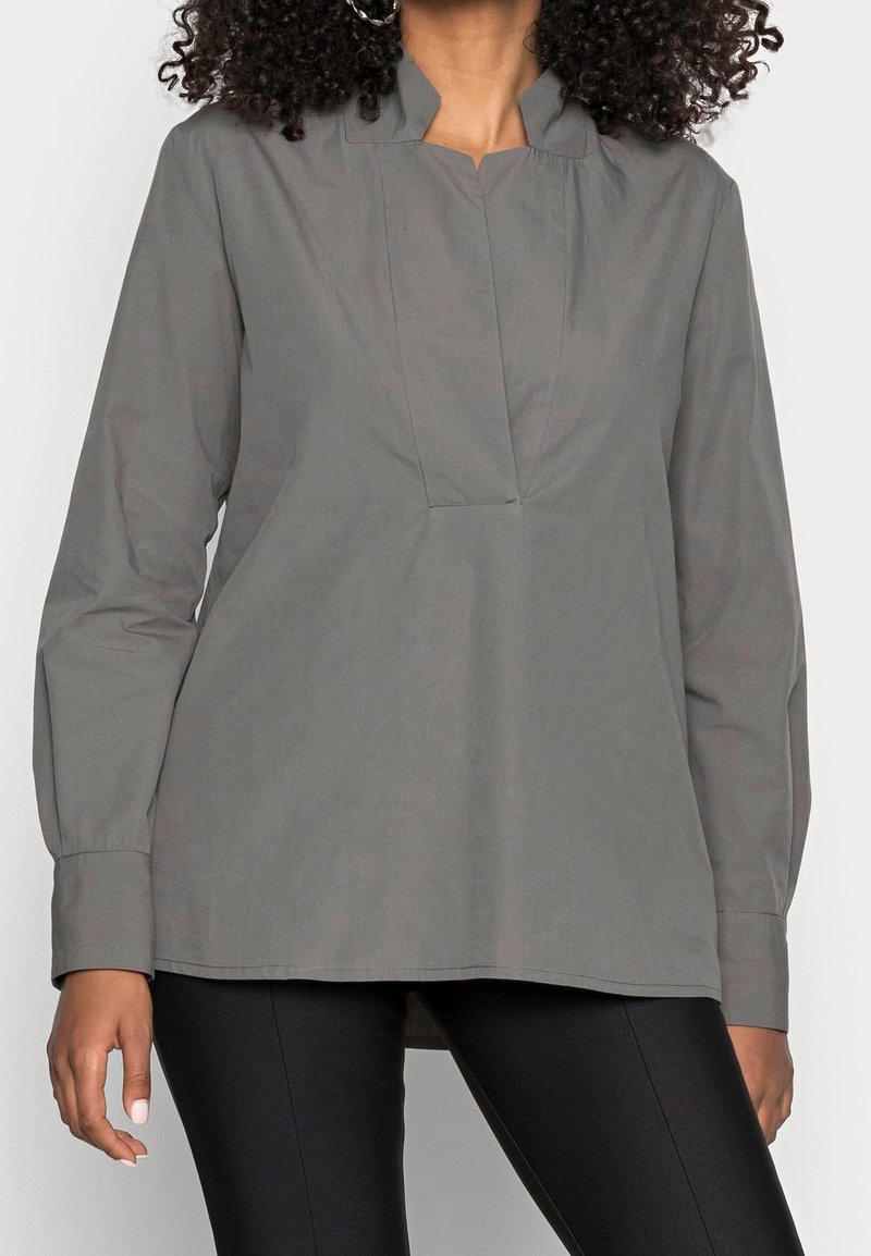 Blusa grigia a maniche lunghe con colletto alto, caratterizzata da una piega frontale e un taglio dritto. Realizzata in un tessuto liscio con una leggera lucentezza.