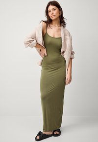 Olijfgroene aansluitende maxi-jurk, lichte stof, spaghettibandjes, gelaagd met een lichtbeige linnen shirt. Zwarte instapsandalen met strikken.