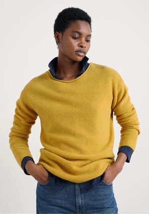 Suéter amarillo de punto con un acabado texturizado, con un cuello de mezclilla por debajo. Combinado con jeans azules, mostrando un ajuste relajado.