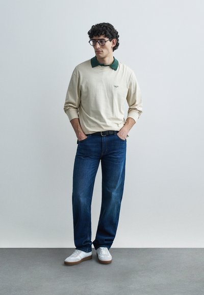 Krēmkrāsains garroku polos krekls ar zaļu apkakli, tumši zili džinsi, balti sporta apavi ar brūnām detaļām; modelis stāv vaļīgi, rokās ieliktas kabatās.