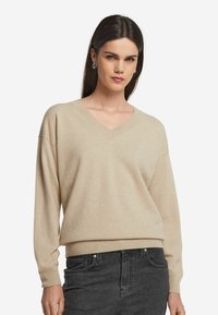 Beige V-Ausschnitt-Pullover aus weichem Strickstoff. Mit überschnittenen Schultern und geripptem Saum. Kombiniert mit dunklen Jeans.