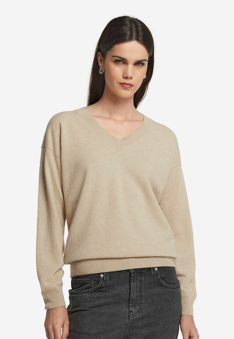 Beige V-Ausschnitt-Pullover aus weichem Strickstoff. Mit überschnittenen Schultern und geripptem Saum. Kombiniert mit dunklen Jeans.