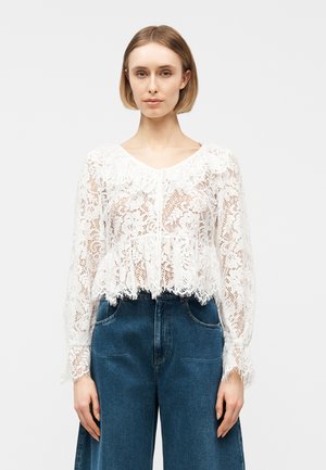 Femme portant un chemisier blanc à manches longues en dentelle florale avec des bords festonnés et un jean bleu à taille haute et jambes larges, debout sur un fond uni.