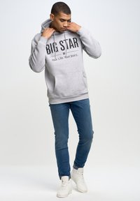 Pelēka kapuce ar priekšējo kengura kabatu, kurai ir "BIG STAR" uzraksts. Apvienota ar ziliem džinsiem un baltām sporta apaviem, fona ir neitrāls.