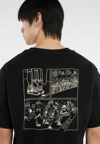 Camiseta de algodón negra con un estampado en estilo caricatura en la parte posterior, que representa a corredores y espectadores en un entorno de carrera con burbujas de diálogo.