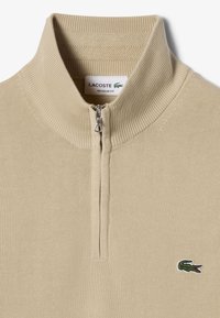 Beżowy sweter zapinany na zamek z prążkowanym kołnierzem i mankietami. Na przodzie ciemnozielone logo Lacoste. Normalny krój, teksturowany materiał dzianinowy.