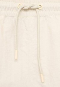 Lys beige shorts med tekstureret stof, der har en snor i taljen i en matchende farve, afsluttet med plastikspidser på snoren.