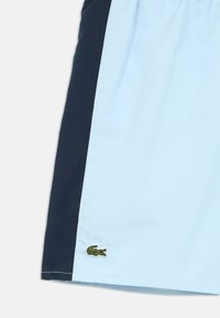 Világoskék rövidnadrág sötétkék oldalsó panelekkel és rugalmas derékpánttal. Kis zöld hímzett krokodil logó a szegély közelében.