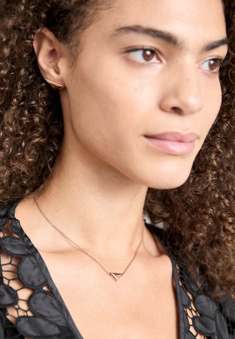 Náhrdelník s trojúhelníkovým přívěskem z růžového zlata na jemném řetízku, nošený modelem s kudrnatými vlasy, na pozadí černého krajkového topu.