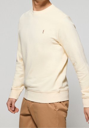 Homme portant un sweat-shirt à manches longues crème avec un petit logo de golfeur brodé et un pantalon marron clair sur un fond neutre.