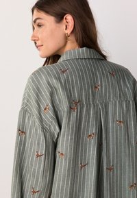Green striped shirt featuring embroidered dog and bird motifs. Collared design with a relaxed fit and buttoned back pleat detail.