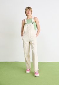 Stripete beige overall med sidelommer, justerbare stropper og logodetalj. Brukt over en grønn topp og sammen med rosa joggesko.