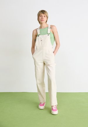 Striped beige overalls with side pockets, adjustable straps, and a logo detail. Worn over a green top and paired with pink sneakers.