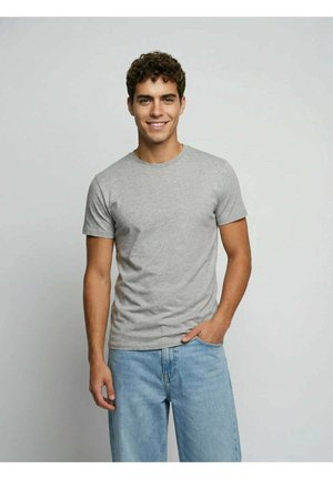 Young man with curly dark hair smiling, wearing a light gray t-shirt and blue jeans, standing against a plain light background.