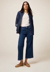 Camisa de mezclilla, camiseta blanca, pantalones vaqueros de pierna ancha azul y mocasines beige. La camisa tiene botones de metal y mangas arremangadas.