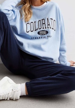 Persona con sudadera de color azul claro con texto "COLORADO 1996 ATHLETIC DEPT", pantalones deportivos azul marino, calcetines blancos y zapatillas blancas, sentada en el suelo.