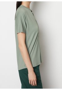 Top corto de color verde claro con cuello henley con botones, ajuste holgado y tejido suave. Llevado con pantalones verdes oscuros, vista de lado.