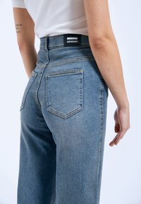 Vaqueros de mezclilla azules con cintura alta, que cuentan con dos bolsillos traseros, costuras amarillas y una etiqueta negra en la cintura. Acabado suave y texturizado.