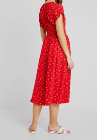 Vestido rojo con flores, de mangas cortas, cintura fruncida, longitud hasta la mitad de la pantorrilla y pequeños patrones de flores blancas. Lleva sandalias blancas tipo deslizar.