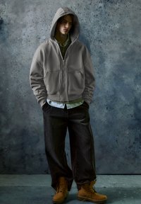 Felpa grigia con zip e cappuccio ampio, abbinata a una maglia a righe e pantaloni larghi marroni. Stivali gialli completano l'outfit su uno sfondo texturizzato.