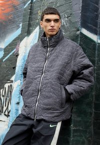 Giacca grigia trapuntata con zip, colletto alto e dettagli neri, indossata sopra pantaloni neri con strisce verdi. Lo sfondo presenta graffiti colorati.