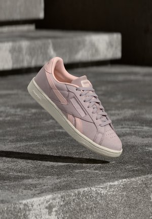 Ανοιχτό ροζ σουέντ αθλητικό παπούτσι Reebok με λευκή σόλα, κορδόνια δεμένα, τοποθετημένο στον αέρα πάνω από μια υφή γκρι σκυροδέματος.