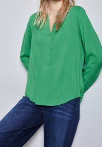 Blusa verde a maniche lunghe con scollatura a V, dettagli con bottoni e spacchi laterali, abbinata a jeans in denim blu scuro. Tessuto dalla texture liscia.