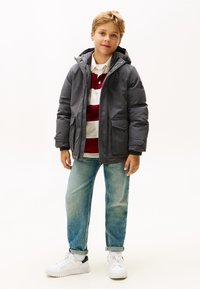 Giacca imbottita grigia con cappuccio, dotata di due tasche frontali. Indossata sopra una polo a righe rosse e bianche e jeans blu chiaro, abbinata a sneakers bianche.