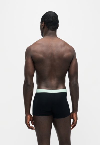 Homme à la peau foncée et aux cheveux courts, debout de dos, portant un boxer noir avec une ceinture claire sur un fond blanc uni.