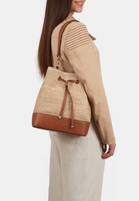 Naturlig vævet stråbucket taske med en brun læderbund, snørelukning og en tan skulderrem, vist på en model iført en beige jakke.