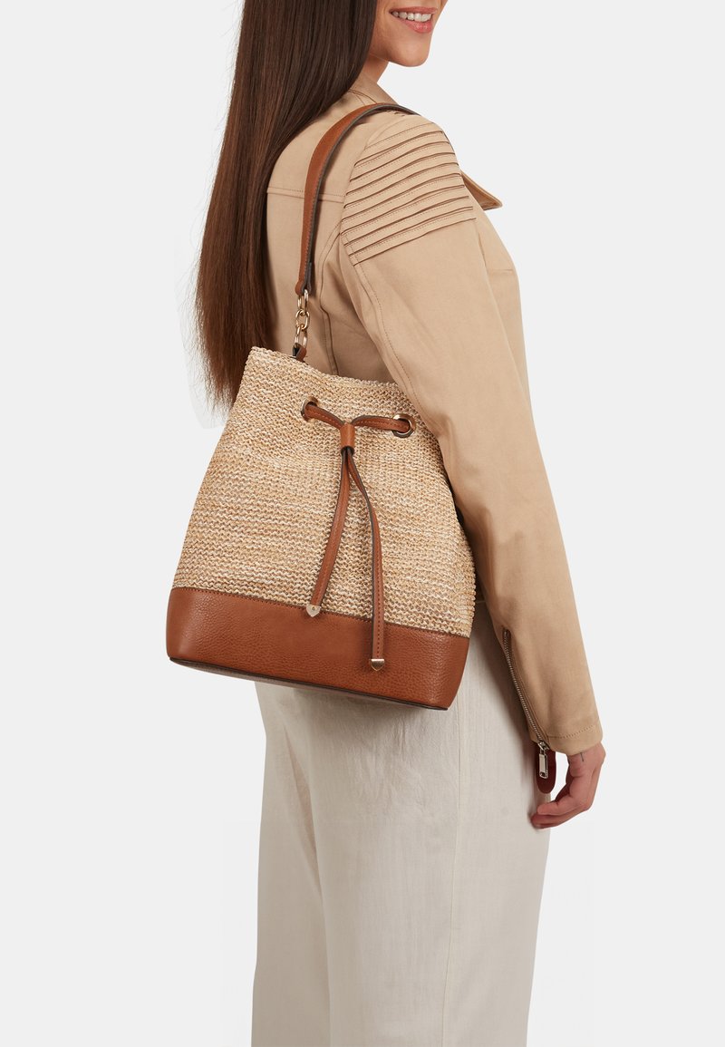 Naturlig vævet stråbucket taske med en brun læderbund, snørelukning og en tan skulderrem, vist på en model iført en beige jakke.