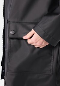 Zwarte waterdichte jas met een grote voorzak met knoopsluiting, een gladde textuur en contrasterende stikdetails.
